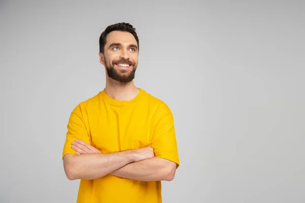 Радостный бородатый мужчина в желтой футболке, стоящий со скрещенными руками и смотрящий в сторону, изолированный на сером — стоковое фото