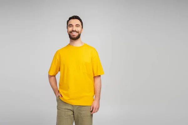 Happy bearded man in yellow t-shirt standing with hand in pocket of beige trousers isolated on grey — Stock Photo
