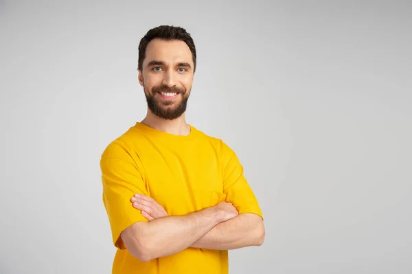Hombre barbudo feliz en camiseta amarilla sonriendo a la cámara mientras está de pie con los brazos cruzados aislados en gris - foto de stock