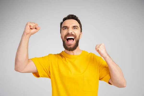 Hombre emocionado en camiseta amarilla gritando y mostrando gesto de triunfo aislado en gris - foto de stock