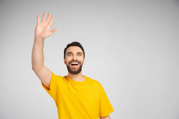 Homem barbudo alegre em t-shirt amarela acenando mão e olhando para longe isolado em cinza — Fotografia de Stock