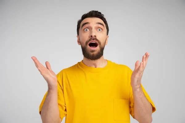 Amazed bearded man in yellow t-shirt standing with open mouth and showing wow gesture isolated on grey — Stock Photo