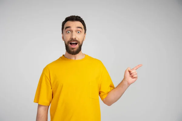 Здивований чоловік у жовтій футболці дивиться на камеру і вказує пальцем ізольовано на сірому — стокове фото