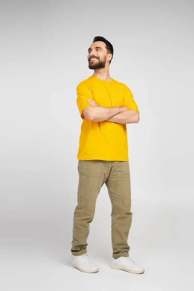 Piena lunghezza di uomo felice in pantaloni beige e t-shirt gialla in piedi con le braccia incrociate e guardando lontano sul grigio — Foto stock