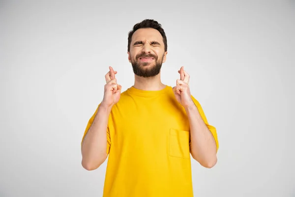Обеспокоенный мужчина в желтой футболке стоит с закрытыми глазами и скрещенными пальцами изолированы на сером — стоковое фото