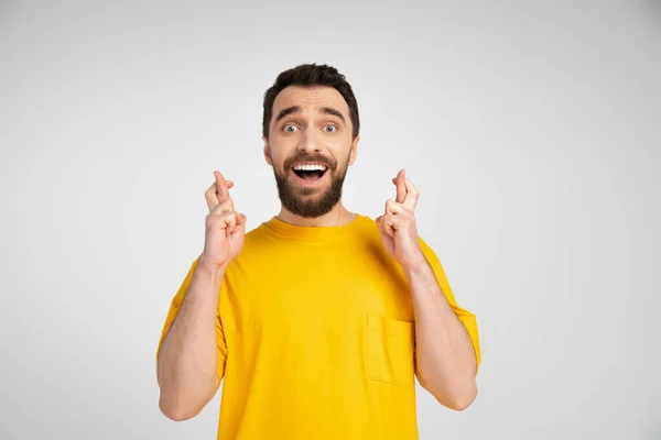 Nervous man in yellow t-shirt holding crossed fingers for luck isolated on grey — Stock Photo