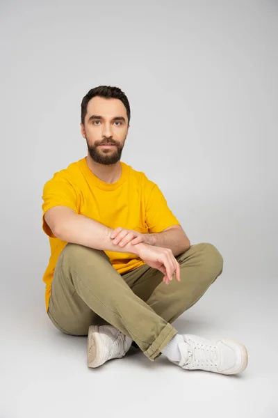 Longueur totale de l'homme en t-shirt jaune et pantalon beige assis avec jambes croisées sur fond gris — Photo de stock