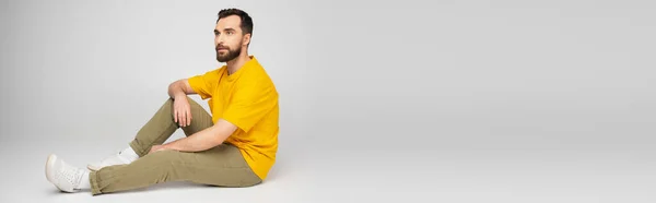 Longitud completa del hombre barbudo en pantalones beige y camiseta amarilla sentado sobre fondo gris, bandera - foto de stock