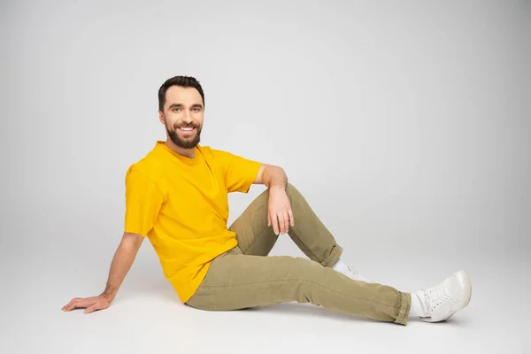 Full length of happy man in beige pants and yellow t-shirt sitting on grey background — Stock Photo