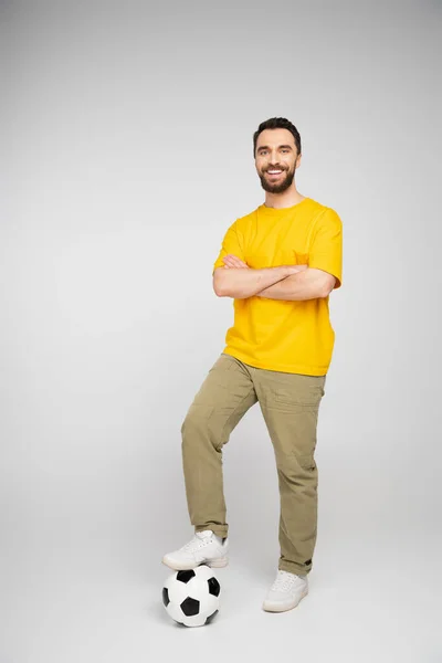 Full length of happy man stepping on soccer ball while standing with crossed arms on grey background — Stock Photo