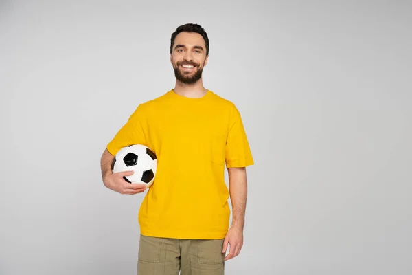 Happy bearded man in yellow t-shirt standing with soccer ball and looking at camera isolated on grey — Stock Photo
