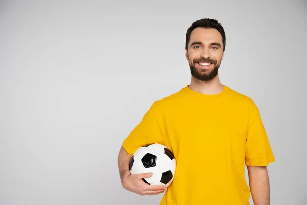Tifoso sportivo sorridente in t-shirt gialla che tiene il pallone da calcio e guarda la fotocamera isolata sul grigio — Foto stock