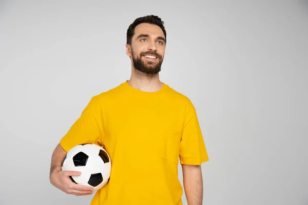 Fanático del fútbol barbudo feliz sosteniendo pelota de fútbol y mirando hacia otro lado aislado en gris - foto de stock