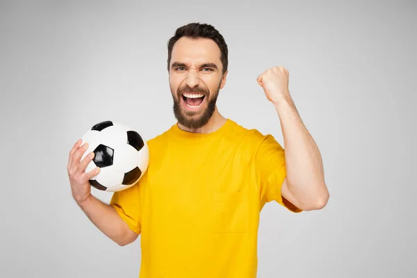 Emocionado fanático de los deportes barbudos con pelota de fútbol gritando y mostrando gesto de victoria aislado en gris - foto de stock