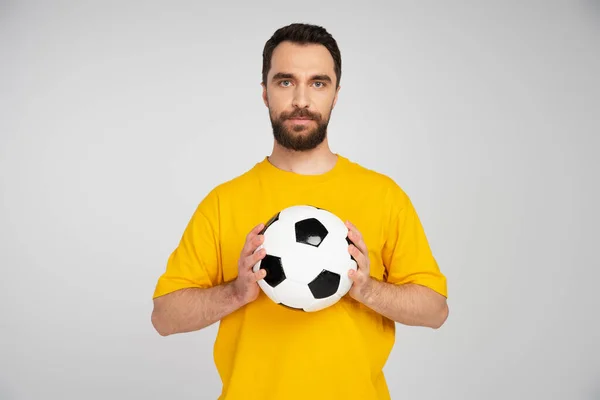 Abanico de deportes barbudos en camiseta amarilla sosteniendo pelota de fútbol mientras mira la cámara aislada en gris — Stock Photo