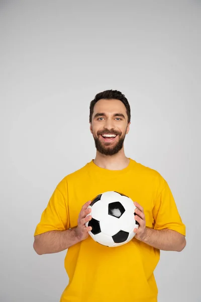 Веселий бородатий спортивний вентилятор з футбольним м'ячем дивиться на камеру ізольовано на сірому — стокове фото
