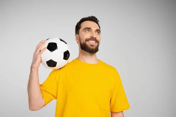 Glücklicher bärtiger Sportfan in gelbem T-Shirt, der einen Fußballball in der Hand hält und isoliert auf grau blickt — Stockfoto