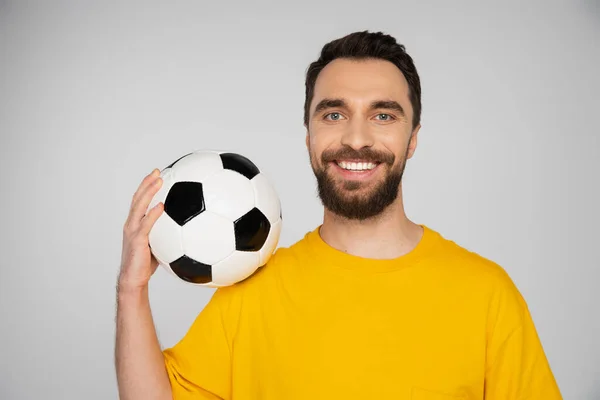 Zufriedener bärtiger Fußballfan in gelbem T-Shirt mit Fußballball und Blick in die Kamera vereinzelt auf grau — Stockfoto