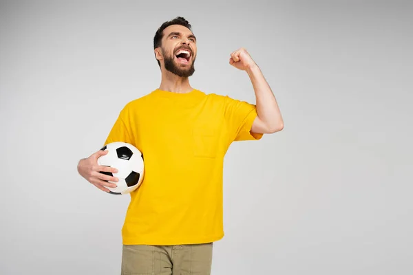 Fanático de los deportes llenos de alegría con pelota de fútbol mirando hacia otro lado y mostrando gesto de victoria mientras grita aislado en gris - foto de stock