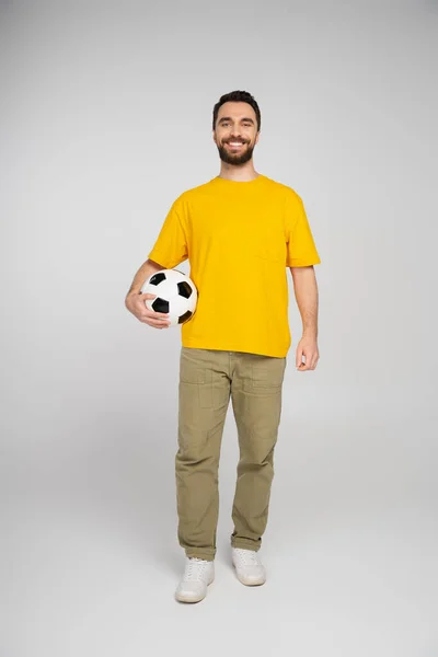 Full length of joyful sports fan in yellow t-shirt and beige pants standing with soccer ball on grey background — Stock Photo