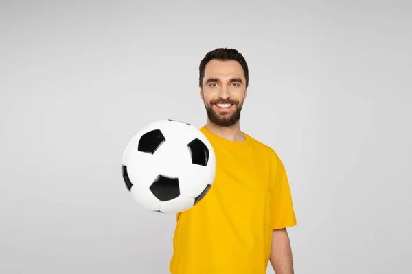 Ventilador de fútbol positivo en camiseta amarilla que muestra la pelota de fútbol en la cámara aislado en gris - foto de stock