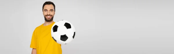 Happy and bearded sports fan showing soccer ball and looking at camera isolated on grey, banner — Stock Photo