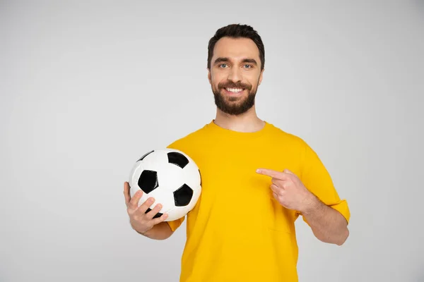 Alegre fanático del fútbol en camiseta amarilla apuntando al balón de fútbol aislado en gris — Stock Photo
