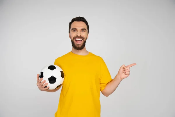 Ventilador de fútbol riéndose de la cámara y apuntando con el dedo mientras sostiene la pelota de fútbol aislado en gris - foto de stock