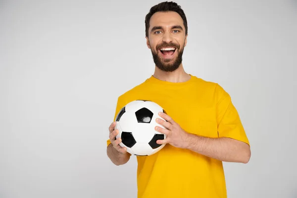 Excited bearded football fan in yellow t-shirt holding ball and laughing at camera isolated on grey — Stock Photo