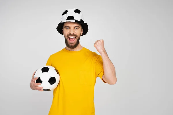 Uomo eccitato in cappello tifoso di calcio tenendo palla e urlando mentre mostra vincere gesto isolato su grigio — Foto stock