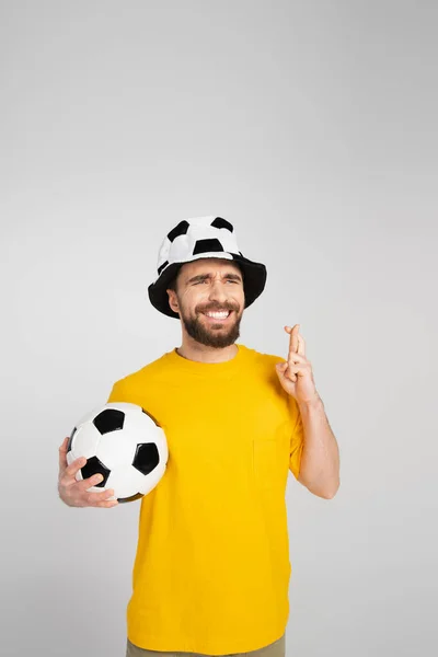 Preocupado ventilador de fútbol en sombrero sosteniendo los dedos cruzados y pelota de fútbol aislado en gris - foto de stock