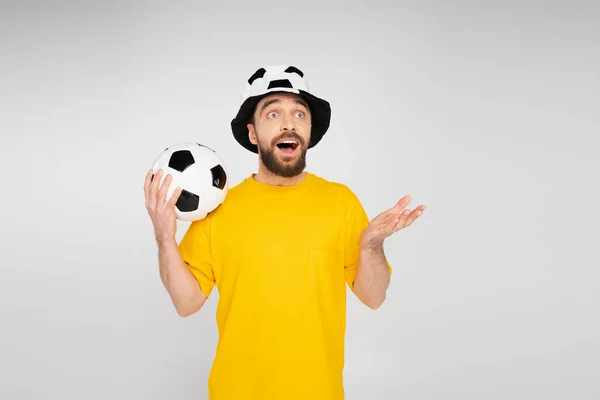 Uomo barbuto stupito in cappello tifoso di calcio che tiene palla da calcio e punta con mano isolata sul grigio — Foto stock