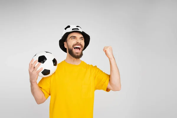 Excited man in football fan hat holding soccer ball and showing triumph gesture isolated on grey — Stock Photo