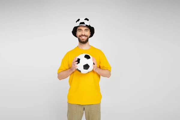 Alegre barbudo hombre en fútbol abanico sombrero celebración fútbol pelota y mirando cámara aislado en gris - foto de stock