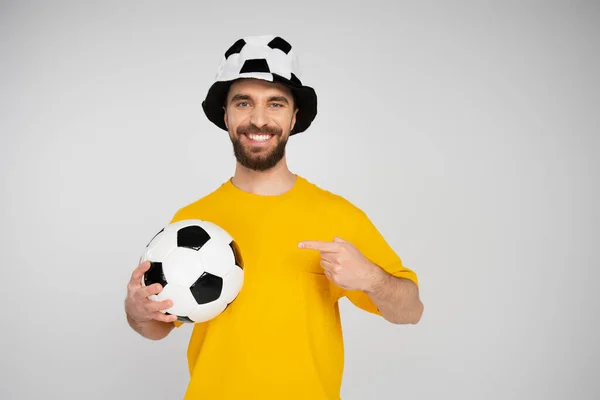 Homem feliz em chapéu de fã de futebol apontando para bola de futebol enquanto olha para a câmera isolada no cinza — Fotografia de Stock