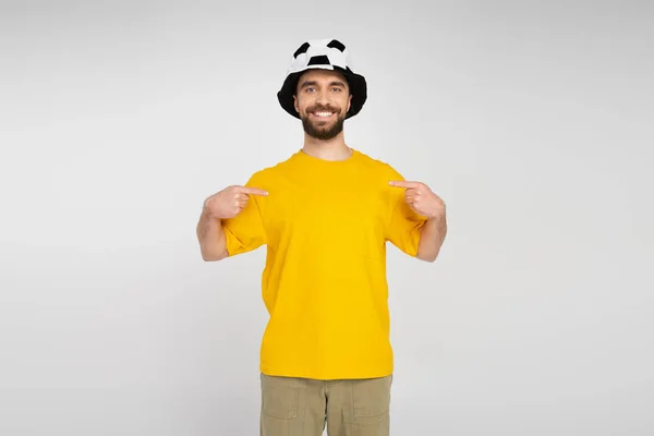 Cheerful bearded man in football fan hat and yellow t-shirt pointing with fingers at himself isolated on grey — Stock Photo