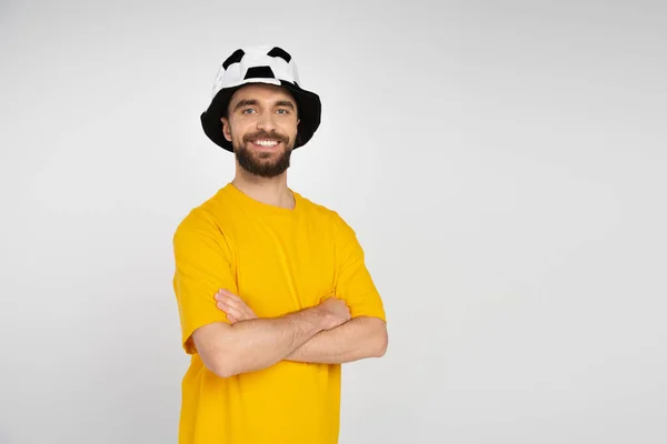 Happy bearded man in football fan hat and yellow t-shirt standing with crossed arms isolated on grey — Stock Photo