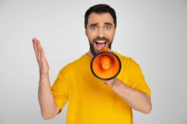 Displeased man in yellow t-shirt shouting in loudspeaker isolated on grey — Stock Photo