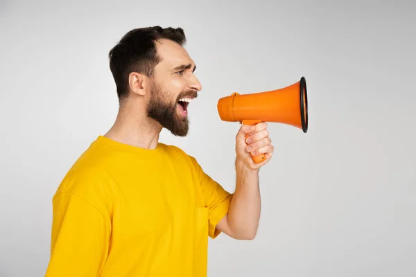 Side view of irritated man in yellow t-shirt shouting in megaphone isolated on grey — Stock Photo