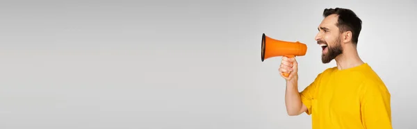 Vue latérale de l'homme barbu mécontent criant en mégaphone isolé sur gris, bannière — Photo de stock