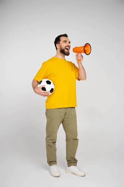 Longitud completa de fanático de los deportes emocionados sosteniendo pelota de fútbol y gritando en megáfono sobre fondo gris - foto de stock