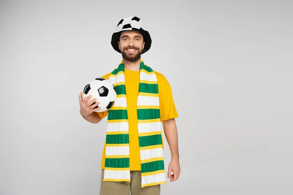 Весёлый футбольный фанат в шляпе и полосатом шарфе, держащий мяч и смотрящий на камеру, изолированную на сером — стоковое фото