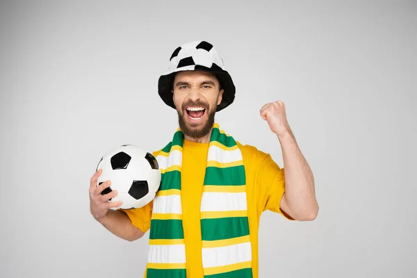 Fanático de los deportes gritando en sombrero y bufanda sosteniendo pelota de fútbol y mostrando gesto de victoria aislado en gris - foto de stock