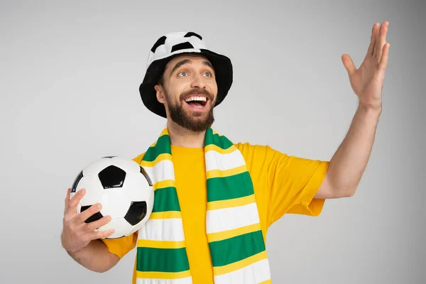 Счастливый и удивленный футбольный болельщик в шляпе и шарфе, держащий мяч, уединившись на сером фоне — стоковое фото