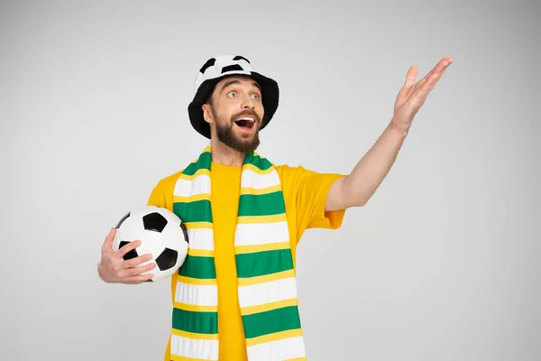 Admirador de los deportes asombrado y alegre en sombrero y bufanda sosteniendo pelota de fútbol y señalando con la mano aislada en gris - foto de stock