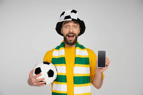 Изумленный футбольный болельщик, держащий футбольный мяч и смартфон с пустым экраном, изолированным на сером — стоковое фото