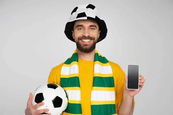 Fanático del fútbol feliz sosteniendo la pelota de fútbol y mostrando el teléfono inteligente con pantalla en blanco aislado en gris - foto de stock