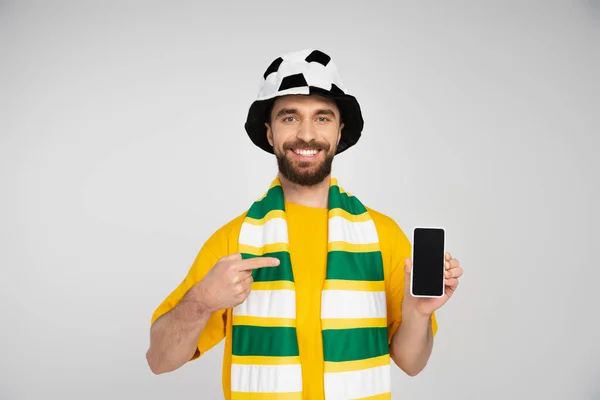Happy bearded man in football fan hat pointing at smartphone with blank screen isolated on grey — Stock Photo