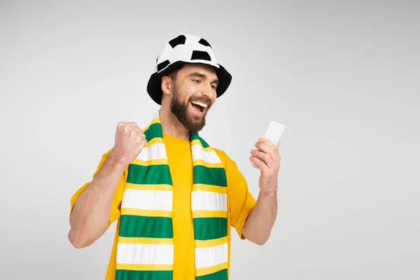 Hombre emocionado en el sombrero del ventilador de fútbol y bufanda rayada mostrando gesto de victoria mientras ve el partido en el teléfono inteligente aislado en gris - foto de stock