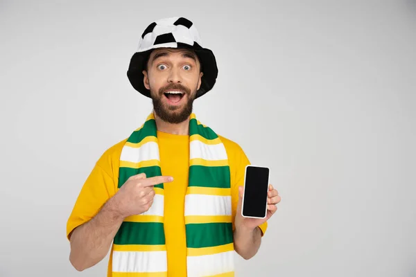 Erstaunter Fußballfan mit offenem Mund, der in die Kamera schaut und auf ein Handy mit leerem Bildschirm in grau zeigt — Stockfoto